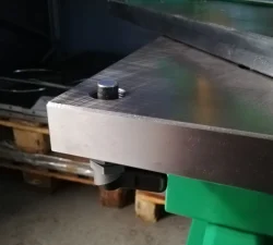 Detailaufnahme-Sicherungspin an einer metallischen Werkzeugplatte, daneben grüne Maschinenkonstruktion und dunkler Werkstattbereich mit Paletten.
