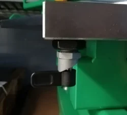Ein schwarzer Sicherungspin sitzt unter einer Metallplatte an einem grünen Werkzeugöffner und fixiert die bewegliche Konstruktion oben.