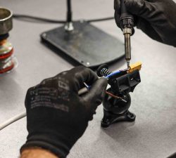 Mit schwarzen Handschuhen wird eine kleine elektrische Verbindung mit dem Lötkolben an einem Bauteil auf dem Tisch verlötet.